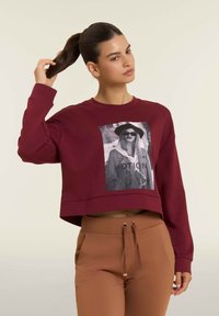 Donna con capelli scuri raccolti in una coda di cavallo che indossa un maglione corto bordeaux con una grafica bianco e nera e pantaloni marroni a vita alta.