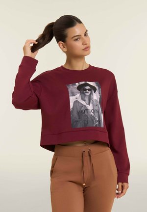 Femme aux cheveux bruns en queue de cheval portant un sweatshirt court bordeaux avec un motif noir et blanc et un pantalon brun taille haute.