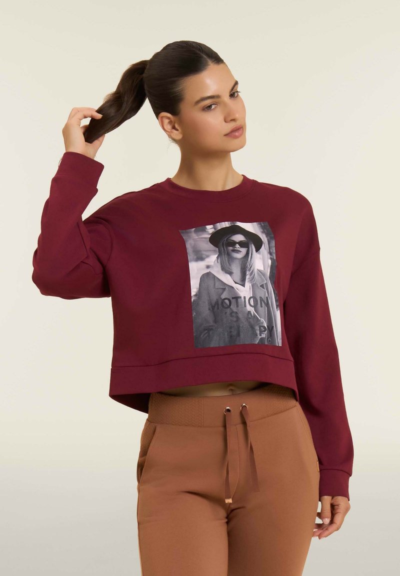 Donna con capelli scuri raccolti in una coda di cavallo che indossa un maglione corto bordeaux con una grafica bianco e nera e pantaloni marroni a vita alta.