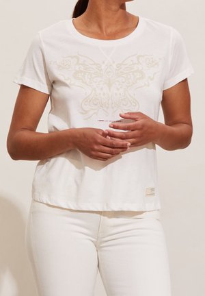 Vrouw draagt een wit shirt met korte mouwen met een licht vlinderontwerp en witte broek, handen samengevouwen ter hoogte van de taille.