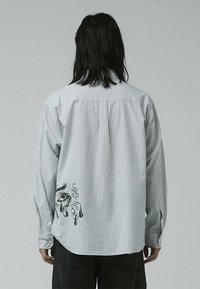 Striped long-sleeve shirt in light gray with black graphic print on the lower left side, featuring a button-down collar and cuffed sleeves.