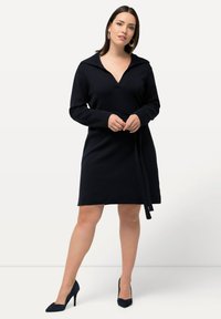 Vestido de punto navy con escote en V y cuello, con mangas largas y cintura ceñida con cinturón, combinado con tacones negros puntiagudos.