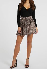 Femme portant un haut noir à manches longues, une mini-jupe à motif animal nouée, et des sandales à talons hauts noires, se tenant devant un fond uni.