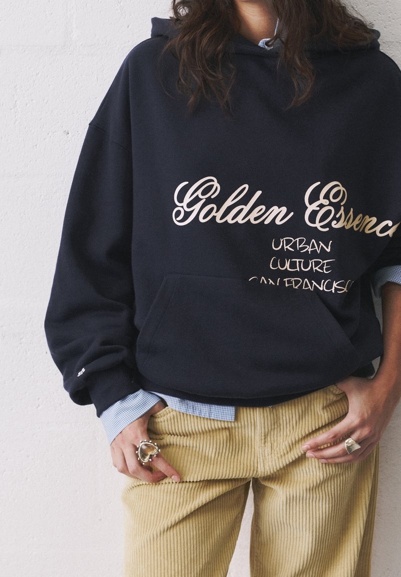 Personne portant un sweat à capuche noir avec le texte « Golden Essence Urban Culture San Francisco », un pantalon en velours beige et des bagues argentées uniques.
