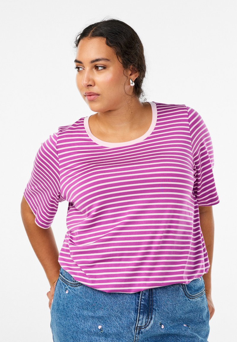 Tume lokkis juustega naine kannab hõbedasi kõrvarõngaid, lilla ja valge triibulist T-särki ning siniseid teksapükse, millel on väikesed tikitud kirsi-mustrid.