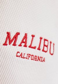 Bílá texturovaná tkanina s červeným vyšitým textem "MALIBU" nad "CALIFORNIA", která má vystouplý, mřížkovaný vzor.