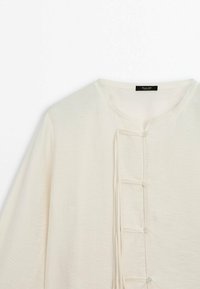 Haut à manches longues de couleur blanc cassé avec encolure ronde et rangée verticale de petits boutons avec boucles en tissu sur le devant.