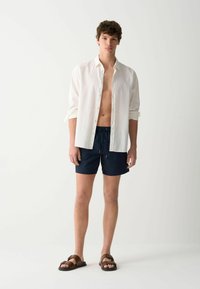 Camicia bianca in lino con bottoni, aperta al collo, abbinata a pantaloni da bagno blu navy. Sandali marroni con cinturini. Sfondo neutro.