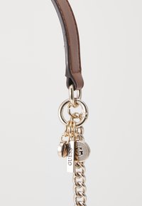 Manico di una borsa in pelle marrone con una catena dorata, anelli per chiavi e etichette metalliche con il marchio "GUESS" e un logo circolare.