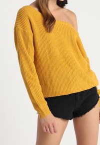 Gul stickad tröja med en axelbandslös design, långa ärmar och texturerad tyg, i kombination med svarta shorts.