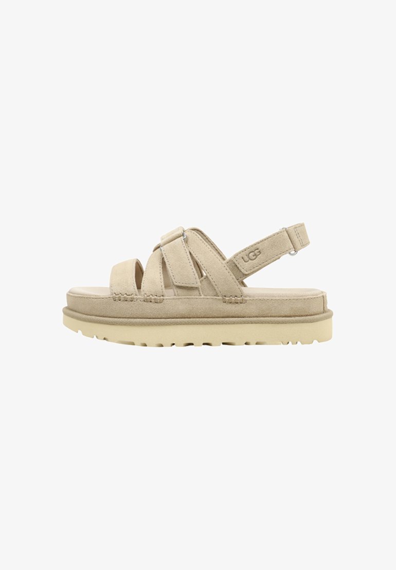 Sandalia de ante beige con tres tiras ajustables, suela gruesa acolchada y logo UGG en la correa del tobillo.