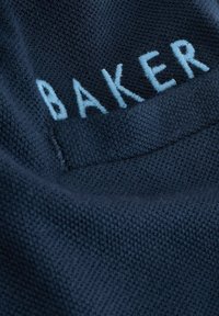 Jūras flotes tekstūra ar gaiši zilu izšūtu "BAKER" tekstu, demonstrējot tīru, mūsdienīgu dizainu ar redzamu šuvju rakstu.