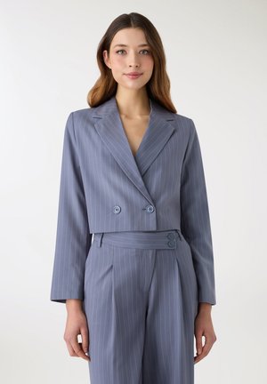 Donna che indossa un blazer corto azzurro a righe sottili con pantaloni a vita alta abbinati su uno sfondo bianco semplice.