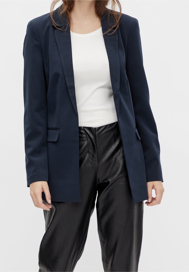 Blazer bleu marine à coupe ajustée, fermeture à un bouton et revers crantés, associé à un haut côtelé blanc et un pantalon en cuir noir.