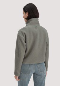 Fleece-Pullover in Hellgrau mit hohem Kragen, strukturiertem Stoff und kleinem Logodetail im Nacken. Kombiniert mit blauen Jeans.