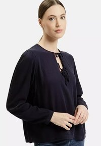 Ciemnoniebieska damska bluzka z długim rękawem, okrągłym dekoltem i detalem związania z przodu, uszyta z teksturowanej tkaniny. Prosty, luźny krój.