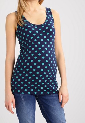 Kobieta ubrana w granatowy bezrękawnik w niebieskie groszki i niebieskie dżinsy, stojąca na jasnoszarym tle.