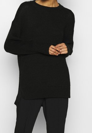 Osoba nosící volný černý pletený svetr s dlouhými rukávy a žebrovanými manžetami, doplněný černými kalhotami na jednobarevném pozadí.