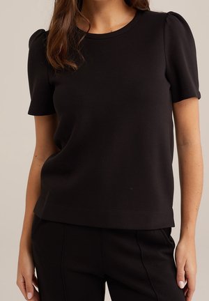 Kvinde iført en ensfarvet sort kortærmet top med let puffærmer og matchende sorte bukser mod en neutral baggrund.