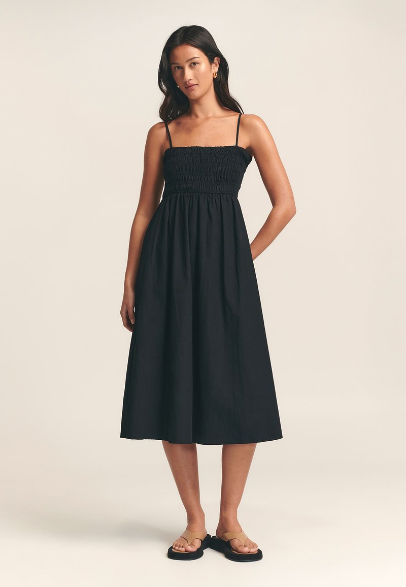 Schwarzes, ärmelloses Kleid mit einem gerafften Oberteil und einem fließenden Rock. Es hat dünne Träger und einen mittellangen Schnitt. Wird mit flachen Sandalen getragen.