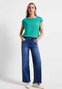 Zelený top s krátkým rukávem spárovaný s volnými modrými džíny. Modelka stojí s rukama v kapsách a má na sobě bílé tenisky. Jednoduchý, ležérní vzhled.