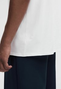 Bílé bavlněné tričko s kulatým lemem, s malým logem na bočním švu. Tkanina působí hladce a lehce.