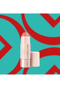 Bâton de blush rétractable avec un dôme rose brillant ; tube blanc avec texte argenté. Sur un fond turquoise avec des motifs géométriques rouges.