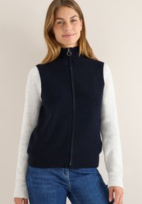 Navy gestrickte Weste mit hohem Kragen und Reißverschluss, kombiniert mit weißen strukturierten Langarmshirts. Glattes, geripptes Gewebe und lässige Passform.
