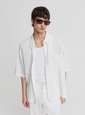 Uomo che indossa una camicia bianca oversize a maniche corte con bottoni, una canotta bianca, pantaloni bianchi e occhiali da sole rettangolari scuri su uno sfondo semplice.