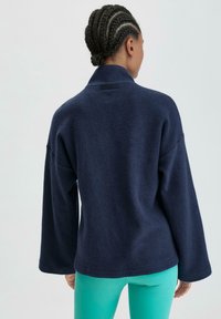 Marineblauer Fleece-Pullover mit hohem Kragen, weiten Ärmeln und texturiertem Finish. Auf der Rückseite befindet sich ein kleiner Markenanhänger. Dazu ein Paar lebhaft türkisfarbener Leggings.