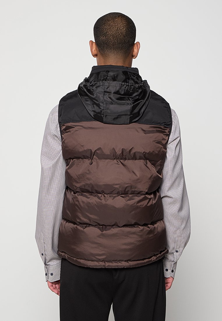 Gilet matelassé marron et noir avec capuche, design matelassé et finition brillante ; porté par-dessus une chemise à carreaux ; présente un col montant.