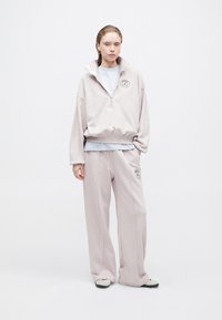 Felpa con zip color rosa chiaro, con colletto alto e logo, abbinata a pantaloni a gamba larga coordinati. Tessuto morbido e vestibilità comoda.