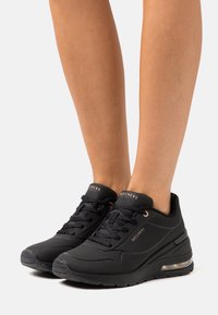 Black Skechers sneakers with air-cushioned sole worn on feet, featuring lace-up front and subtle rose gold branding accents.