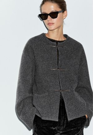 Giacca di lana grigia con colletto rotondo, tre chiusure a toggles e tasche; abbinata a occhiali da sole neri e pantaloni scuri.