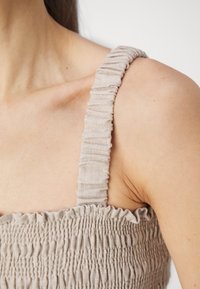 Světle béžový top s šňůrkovými ramínky, vyrobený z texturované látky a s nařasenými detaily, který zvýrazňuje přiléhavý střih.