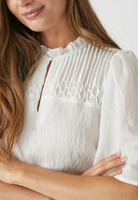 Blusa bianca con colletto alto arricciato, pince verticali e apertura a chiave. Tessuto morbido e testurizzato con maniche corte a sbuffo.