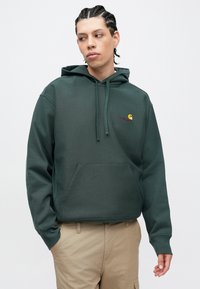 Sudadera con capucha verde oscuro de tela suave, con bolsillo frontal, capucha con cordón y logo circular amarillo con texto rojo en el pecho.