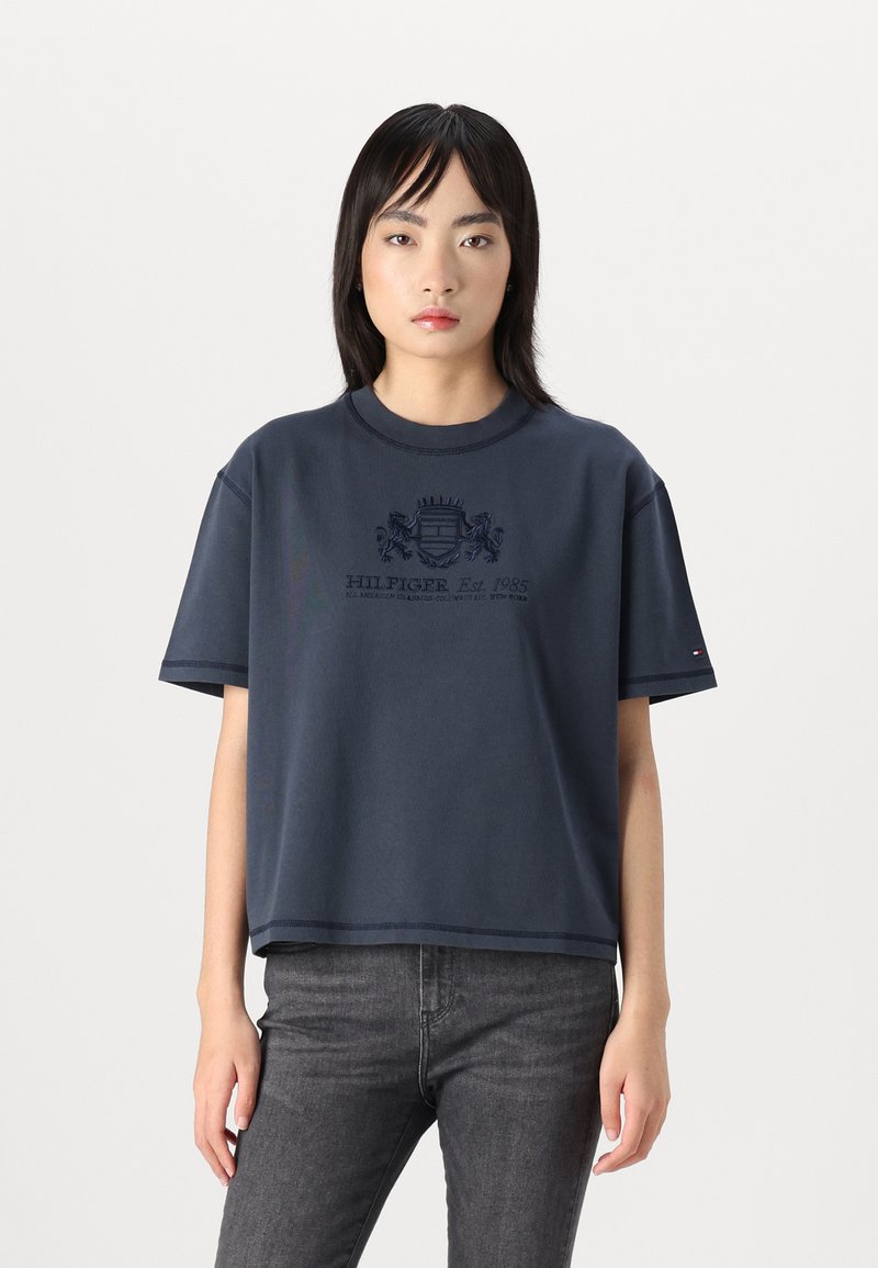 Mørkeblå beskåret t-shirt i blødt stof, der har korte ærmer og et broderet logo med skjolddesign på fronten.