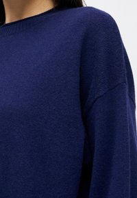 Femme portant un pull en tricot bleu marine avec un col rond côtelé et des épaules tombantes, sur un fond uni.