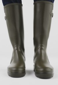 Tummanvihreät kumisaappaat, joissa sileä pinta, korkea malli ja sivulla säädettävä solkikoriste. Tasapohja ja pyöreä kärki.