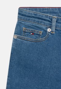 Jeans de denim con tejido azul, que presentan un pequeño bolsillo frontal, costuras doradas y un botón metálico. Destaca un acento rojo cerca del bolsillo.