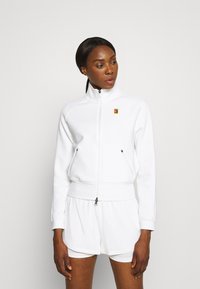 Hvid zip-up jakke med høj krave, sidelommer og logo accent. Glat, struktureret stof; parret med hvide shorts under.