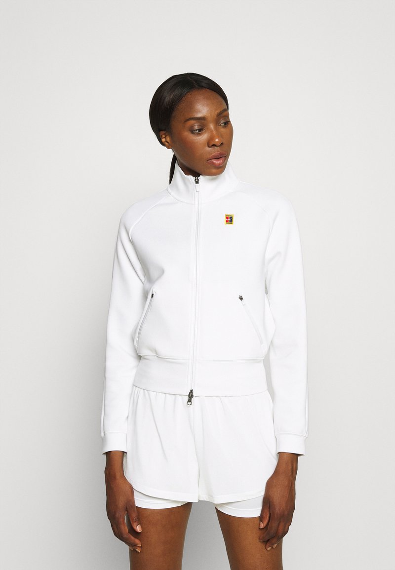 Hvid zip-up jakke med høj krave, sidelommer og logo accent. Glat, struktureret stof; parret med hvide shorts under.