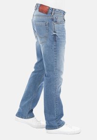 Jeans in denim azzurro con taglio dritto, cinque tasche e cuciture a contrasto. Presentano un'etichetta in pelle marrone sulla cintura. Sneakers bianche.