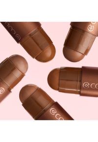 Cinq teintes variées de fond de teint stick crémeux avec des capuchons marron, disposées sur un fond rose pâle.