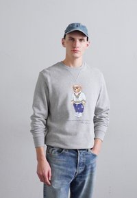 Gray crewneck sweatshirt featuring a colorful bear graphic in sporty attire. Paired with light blue jeans, worn casually.