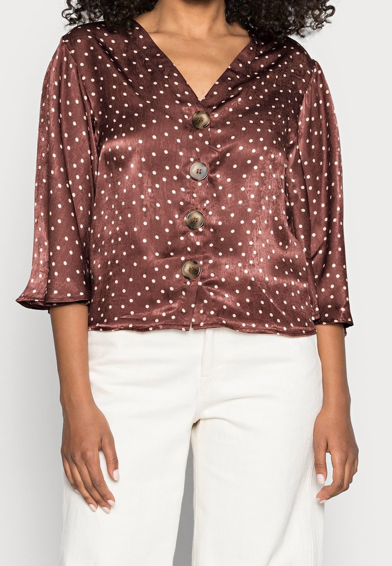 Brun satinblus med vita prickar, V-ringning, trekvartsärmar och fem stora knappar fram. Slät textur.