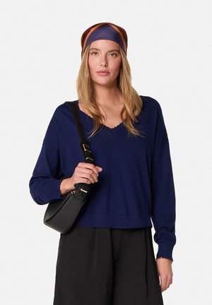 Marineblauwe gebreide trui met een golvende zoom en lange mouwen, gecombineerd met een zwarte crossbody tas en een gedessineerde zijden hoofddoek.