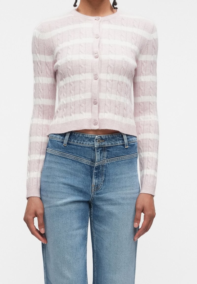 Femme portant un cardigan en maille torsadée court à rayures roses et blanches avec des boutons, associé à un jean bleu clair taille haute.