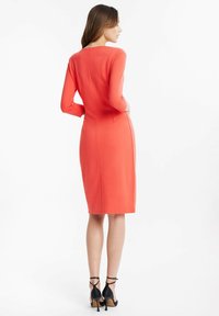Koralfarbenes knielanges Kleid mit langen Ärmeln, figurbetonter Silhouette und Nahtdetails auf der Rückseite. Getragen mit schwarzen High Heels.